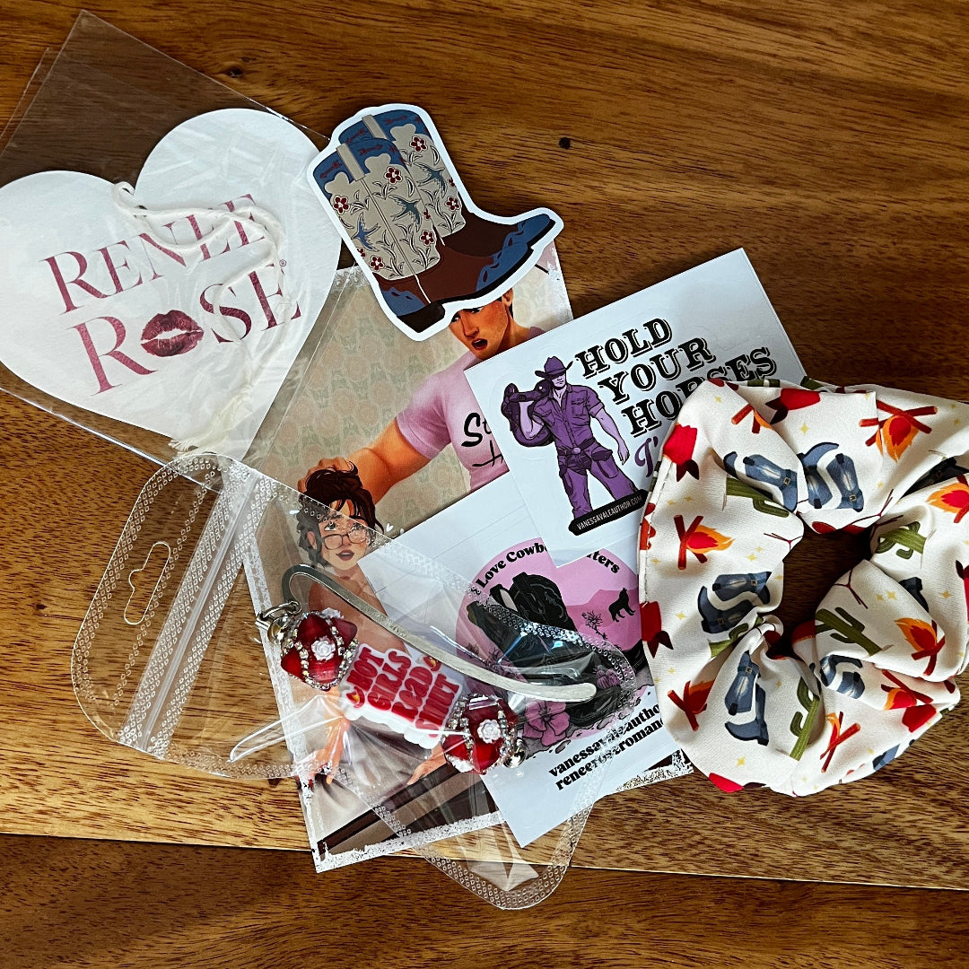 A flat-lay collection of themed merchandise on a wooden tabletop, featuring a white hair scrunchie with a red and multicolor cactus pattern, a white heart-shaped air freshener reading 'Renee Rose', a cowboy boot sticker, and other printed stickers and cards.