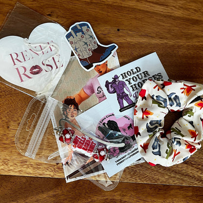 A flat-lay collection of themed merchandise on a wooden tabletop, featuring a white hair scrunchie with a red and multicolor cactus pattern, a white heart-shaped air freshener reading 'Renee Rose', a cowboy boot sticker, and other printed stickers and cards.