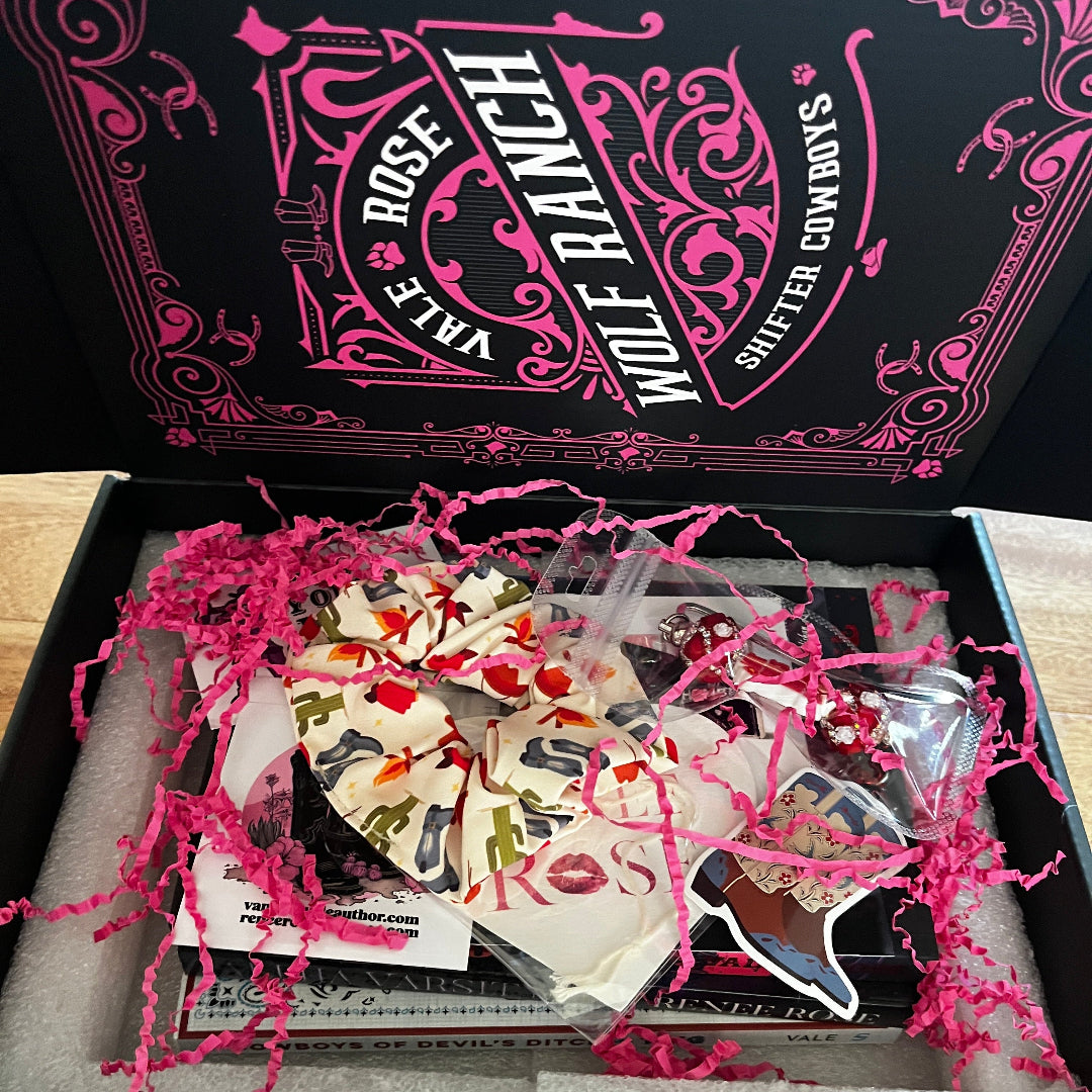 An open Wolf Ranch gift box on a wooden table, showing a variety of books stickers, cards, hair scrunchie and metal and beaded bookmark nestled in pink shredded paper. The black and pink branded box lid is visible in the background.