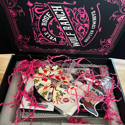 An open Wolf Ranch gift box on a wooden table, showing a variety of books stickers, cards, hair scrunchie and metal and beaded bookmark nestled in pink shredded paper. The black and pink branded box lid is visible in the background.