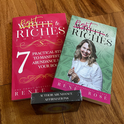 Two author abundance books and a small affirmation box arranged on a wooden floor. The books are the 'Write to Riches' book and accompanying journal by Renee Rose, with one cover in pink and the other in green, and the box in front reading 'Author Abundance Affirmations.