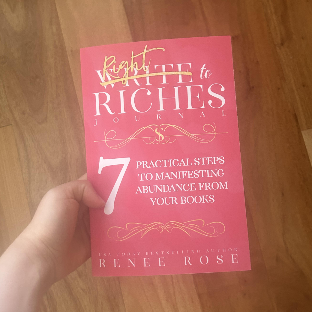 A white person's hand holding a paperback. The book cover's background is pink. In white text is the title "Write to Riches Journal." In yellow, the word "Write" is crossed out are replaced with "Right." Underneath that is a gold dollar sign design. Further down in white text is "7 practical steps to manifesting abundance from your books." On the bottom of the cover in white text is "USA Today Bestselling Author -- Renee Rose"