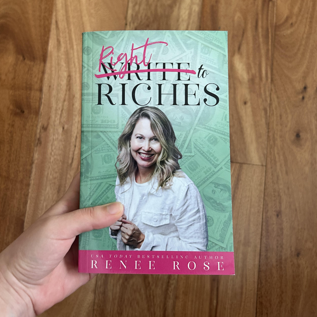 A white person's hand holding a paperback. The book cover's background is green and features $100 bills. It features a women -- Renee Rose-- in white white shirt and jacket smiling out. In black text is the title "Write to Riches." In pink, the word "Write" is crossed out and replaced with "Right." On the bottom of the cover in white text is "USA Today Bestselling Author -- Renee Rose"