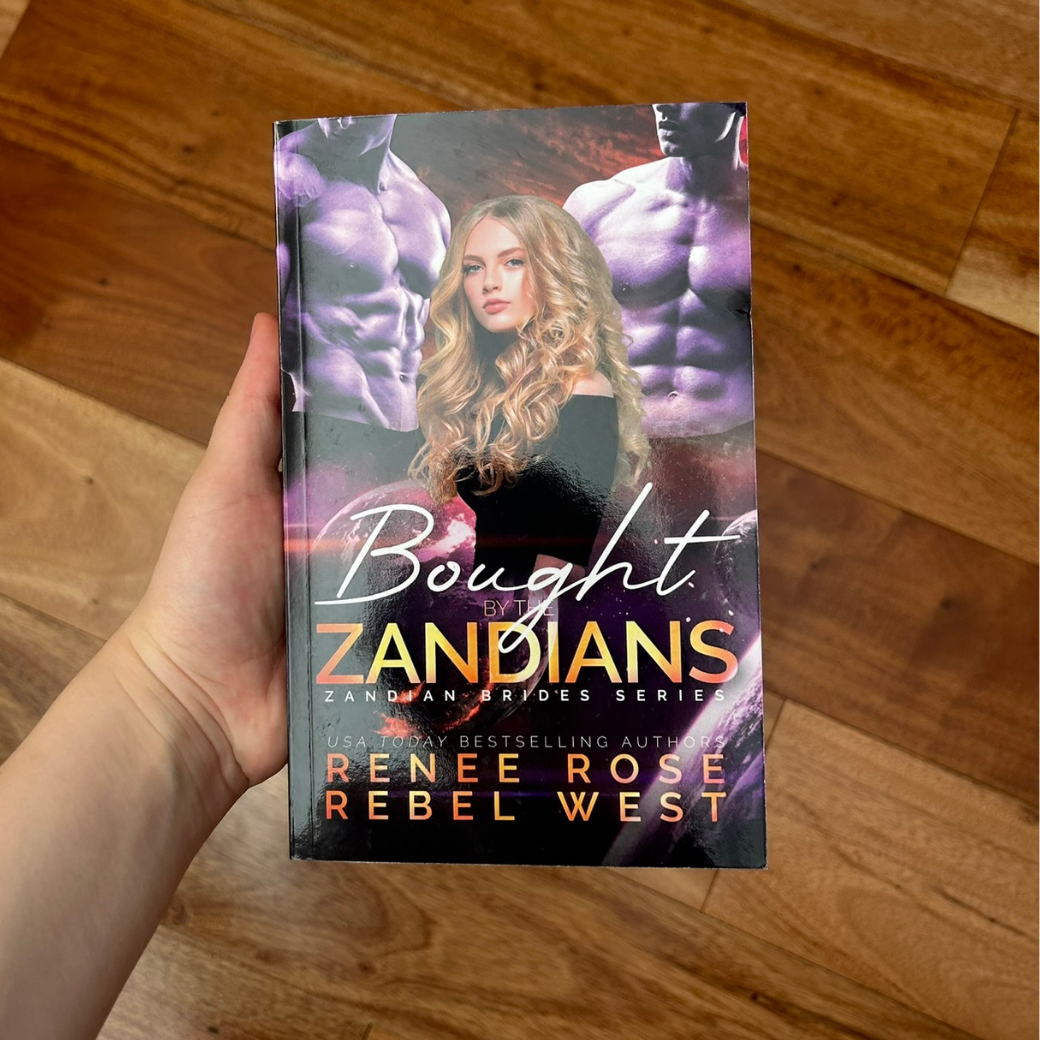 A white person's hand holding a paperback. The book cover features a blonde women with curled hair wearing a tight black shirt. There are two buff shirtless men behind her. The background is stars and planets. In white and gold text is the title "Bought by the Zandians Zandian Brides Series." Underneath that is "USA Today Bestselling Authors--Renee Rose Rebel West"