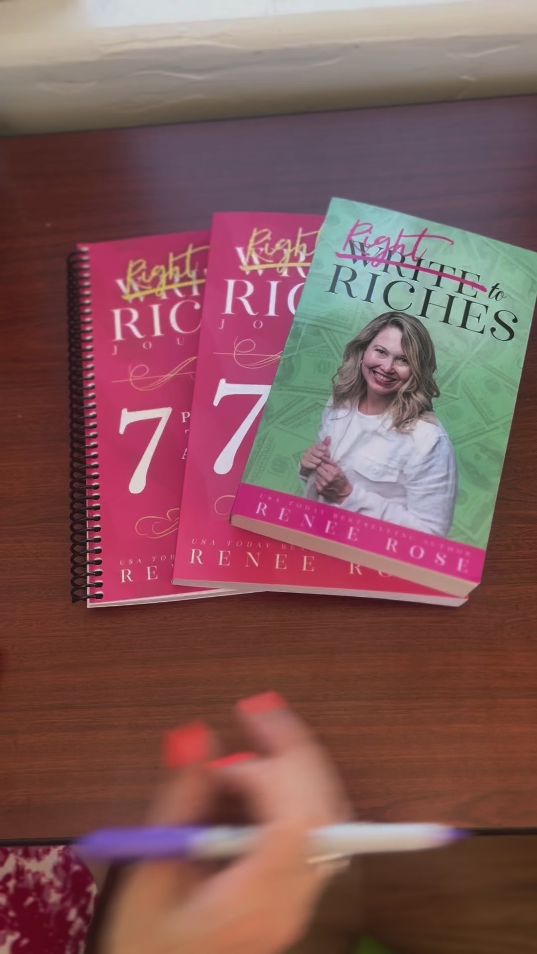 Sped up video. The Write to Riches book, and two versions of the journal are fanned out. A white woman -- Renee Rose -- opens the book, and signs it "ask, believe receive! Renee Rose"