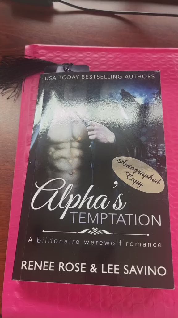 Video of a white woman -- Renee Rose -- showing off a paperback of "Alpha's Temptation." She pulls out stickers and bookmarks out of the book and shows that hand written in a thin purple sharpie is "Love in the elevator," then signed by Renee Rose and Lee Savino. 